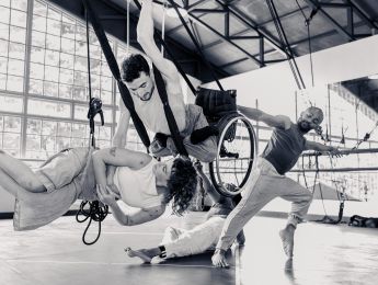 4 performers dance together. One is in a flying wheelchair, one is suspended in a harness. Two others are ground based dancers, interacting with those in the air.