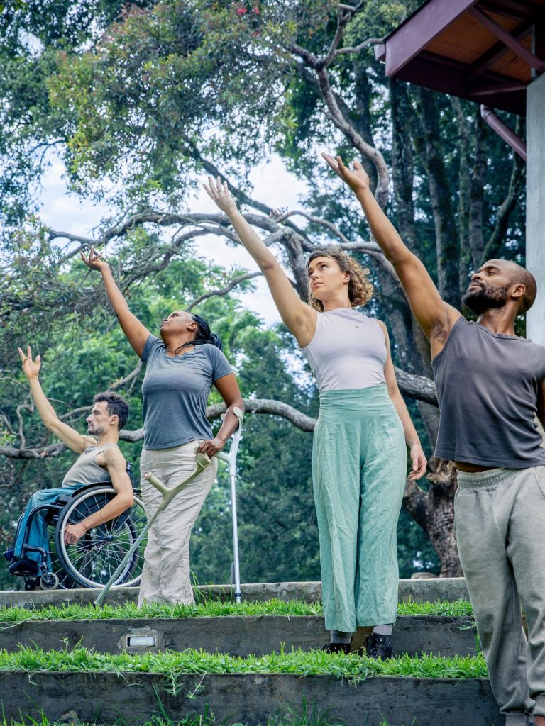 4 Performers stand in a line, all reaching their arm up. They are in a tropical garden, each on a garden step.